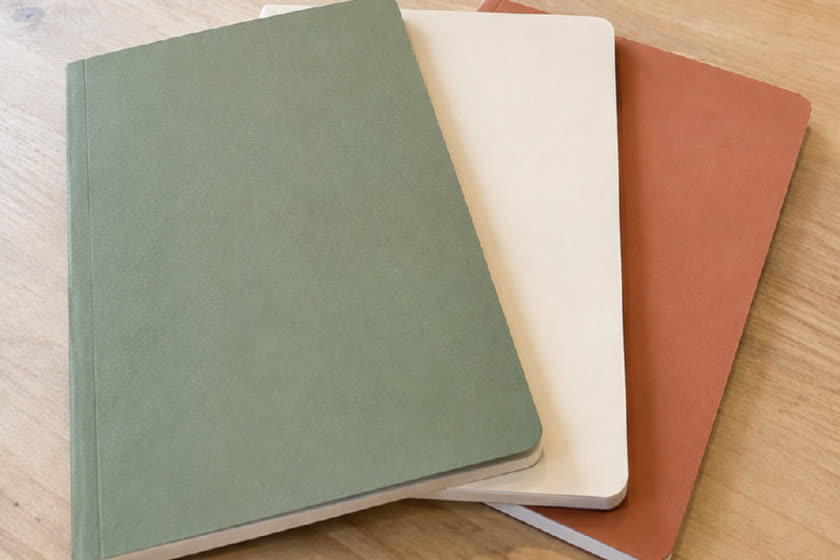 Ahşap masada yelpaze gibi dizilmiş yeşil, krem ve kiremit üç defter